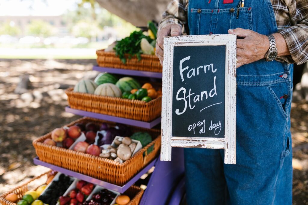 Man in overalls holds farm stand sign with veggies in background